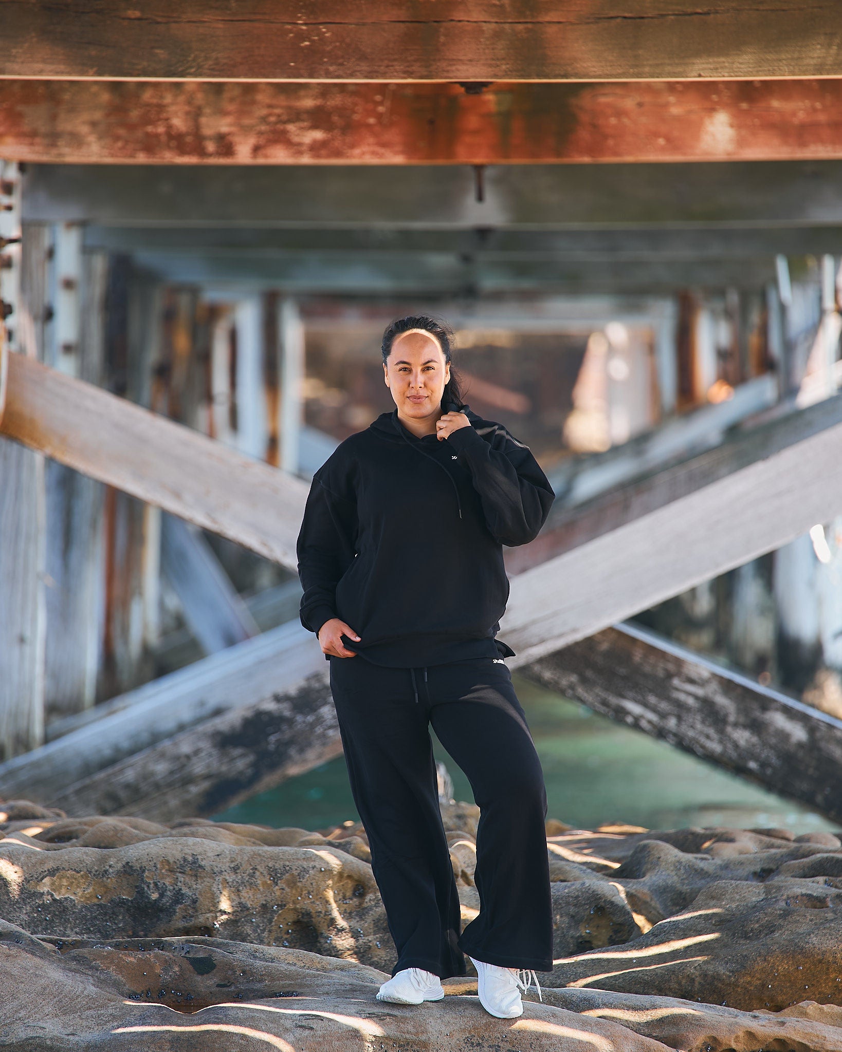 Woman in Womene Lounge Set, black cotton hoodie and joggers, standing on coastal rocks at golden hour