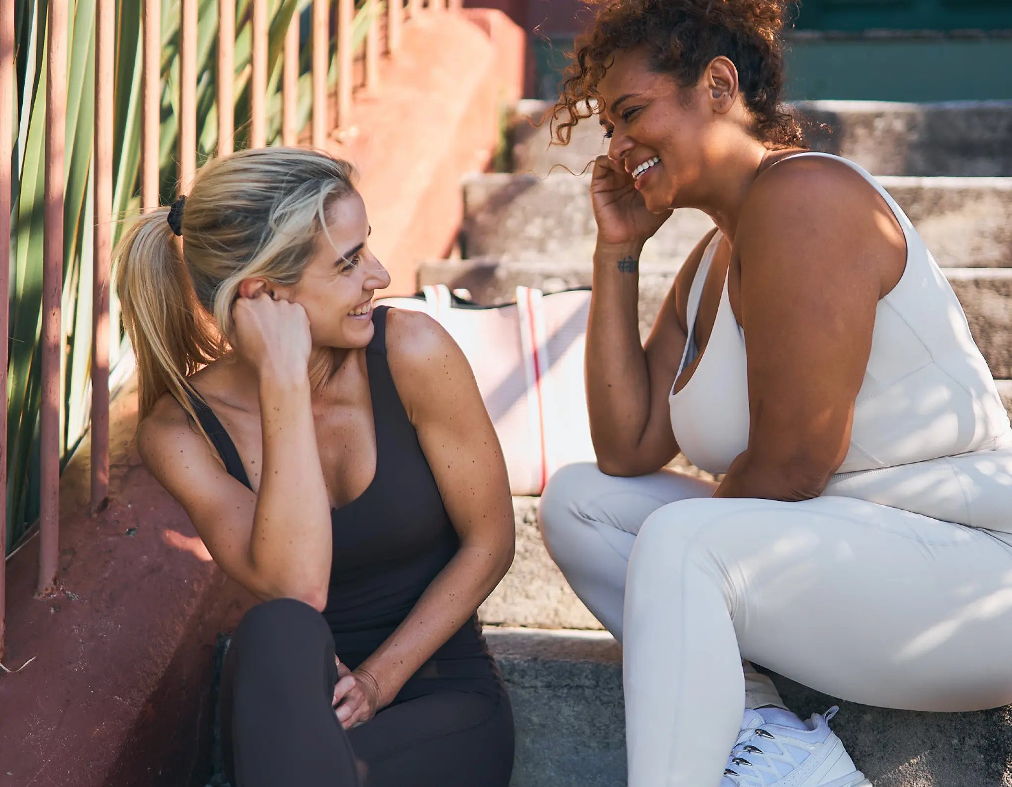 Two women sitting outdoors, one in a black tank top and leggings, the other in a white outfit, both smiling. Womene Australian women's activewear collection