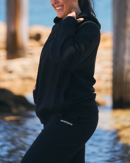 Detail of black cotton hoodie cuff and embroidered Womene mark, 100% cotton fabric
Woman walking in Moments Cozy Hoodie and Joggers, black cotton, Australian coastline
Three-quarter view of Womene Lounge Set in black, hood up, cotton hoodie and matching joggers
