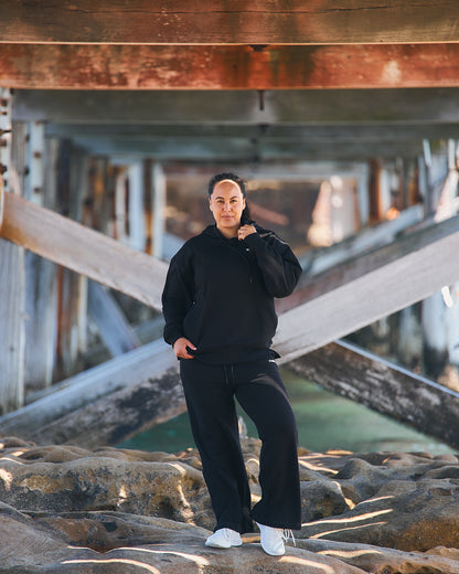 Woman in Womene Lounge Set, black cotton hoodie and joggers, standing on coastal rocks at golden hour
