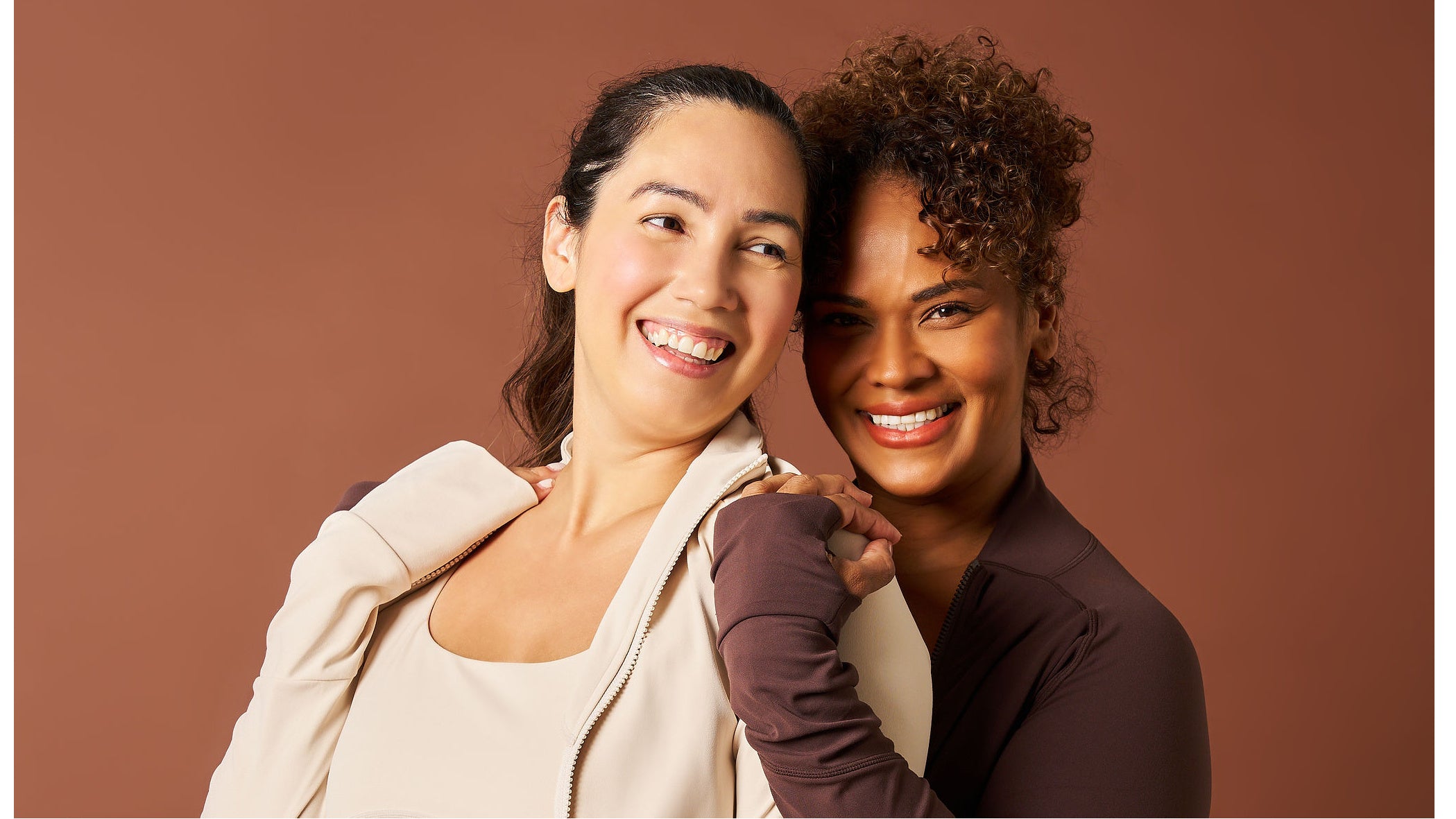 Two women embracing each other against a brown background. Woman wearing Womene Moments activewear collection